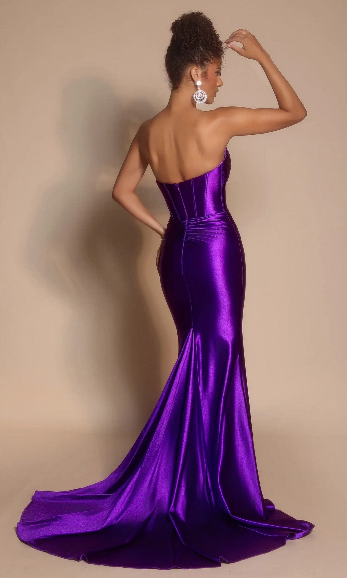 A woman faces away, wearing the Portia and Scarlett XO080 long prom dress with a strapless satin design, floor-length train, and large circular earrings, set against a plain beige background.